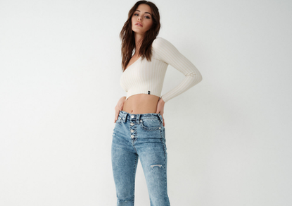 A woman wearing jeans and a top available from True Religion in front of a white background.