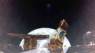 the dark, flat refelctive panels of a space craft line the foreground, with gold-foil wrapped copmonents sticking out in the middle. Behind, the blue round Earth in teh black of space.