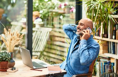 Mature businessman leaning back and smiling using mobile phone, working from home, sitting at desk in front of laptop.