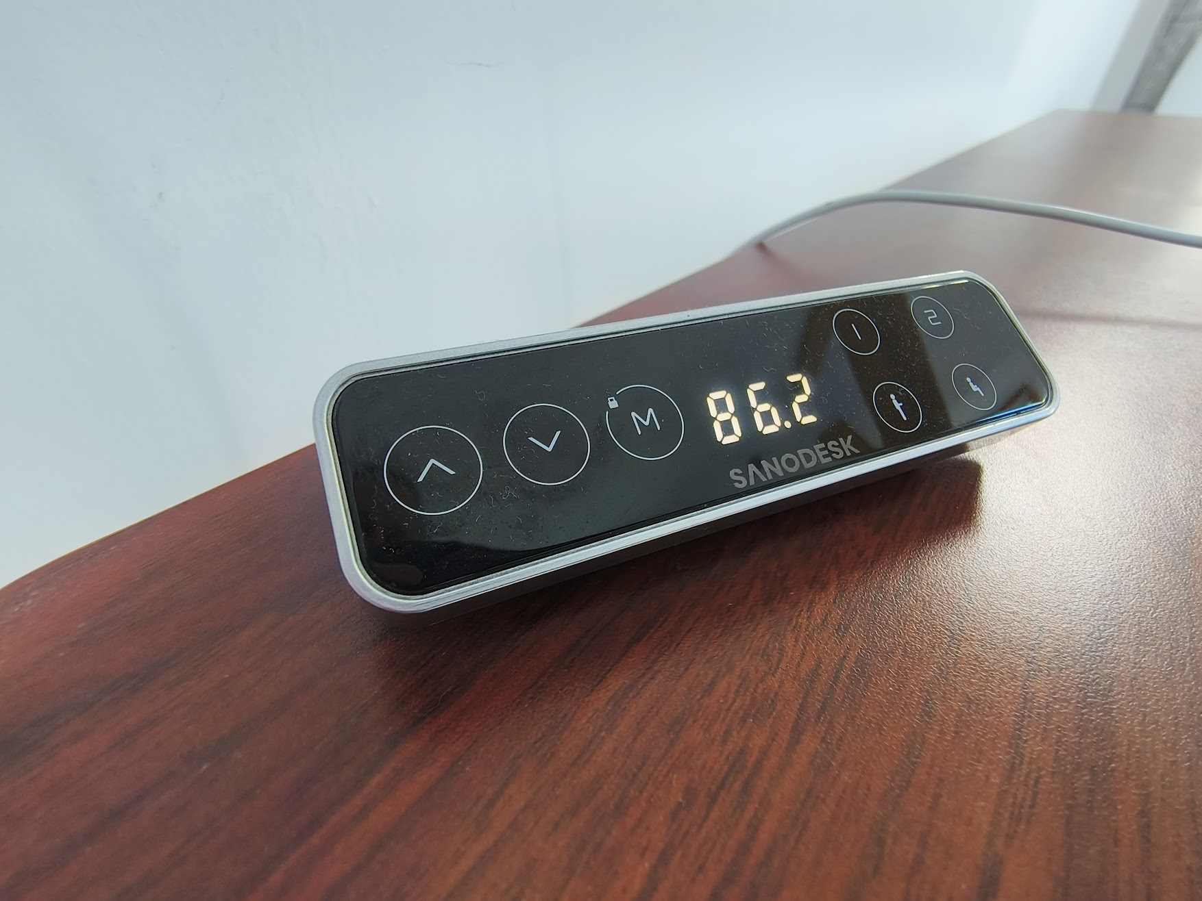 A close-up of a Sanodesk digital control panel with a glowing height display of 86.2 and various touch-sensitive buttons for adjusting a standing desk.