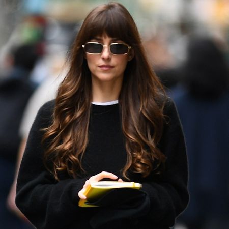 Dakota Johnson walking in New York City wearing a black sweater, dark-wash jeans, and brown Nike Cortez sneakers