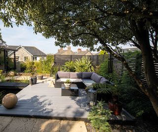 A shaded garden seating area with grey composite decking a mature tree and grey corner sofa