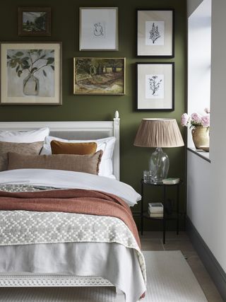 green bedroom with gallery wall, rustic feel, white painted bed, ochre blanket and cushion, glass based lamp, linen cushions, cream rug