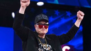 Elon Musk at an event with his hands raised in the air, wearing a cap and reflective sunglasses