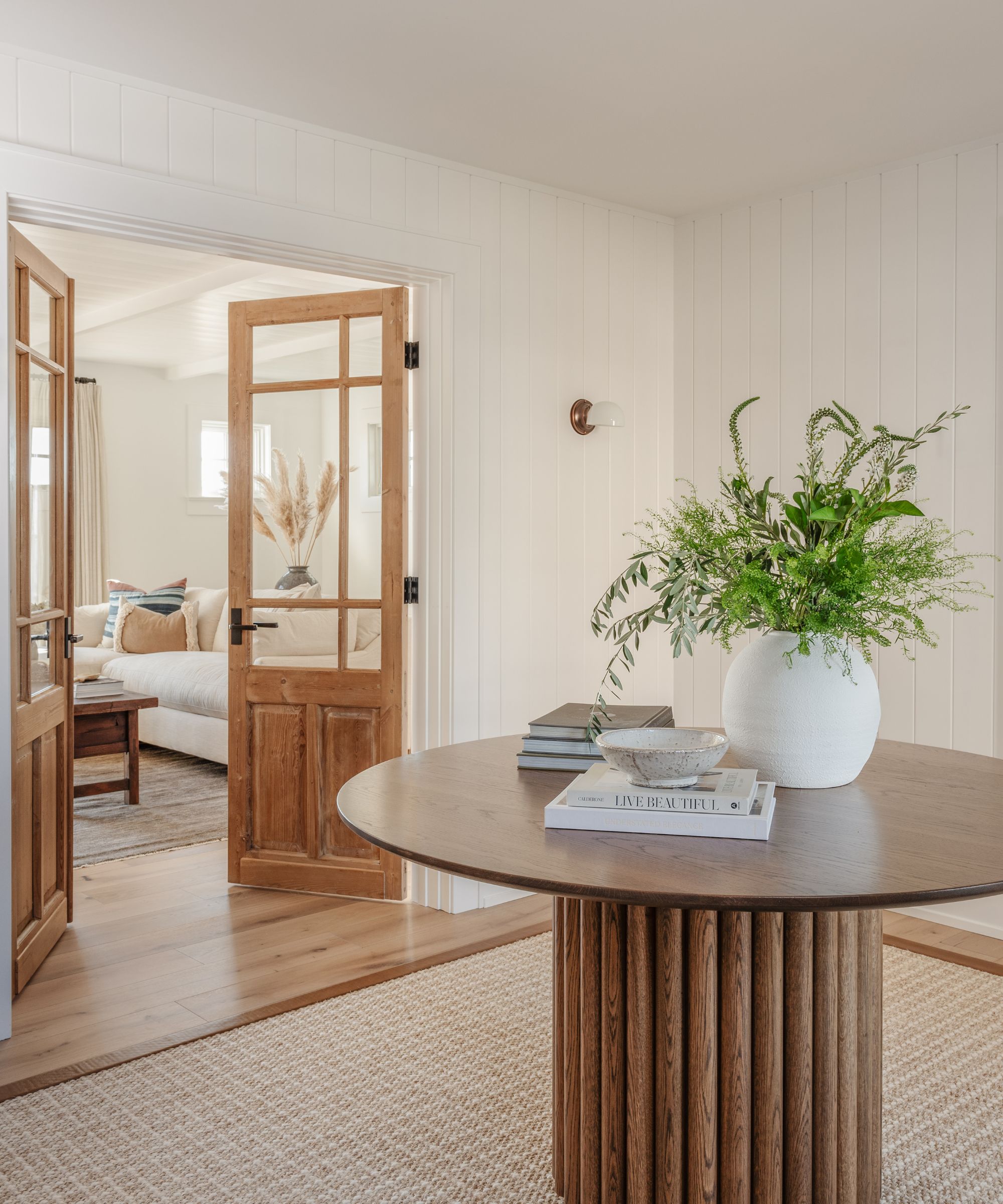 Entryway with panelled white walls, jute rug on wood floor, large wood round table with books, white vase and foliage and bowl and wooden doors