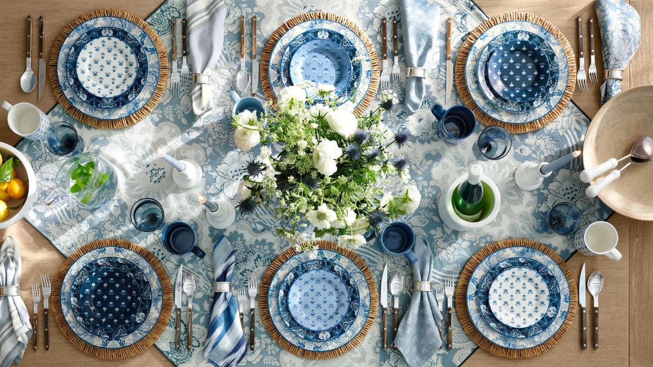 Bird&#039;s eye view of six blue and white dinner plates with metal cutlery on a blue and white tablecloth on a wooden table.