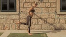 Pilates instructor Rachel Lennon exercises outside on a Pilates mat. She is standing on her right leg, hands on hips, with her left leg bent at the knee and foot pointing back. Behind her we see a stone wall.