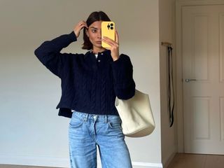  @smythsisters taking a mirror selfie in jeans and a navy sweater, with a cream leather tote and yellow iPhone case.