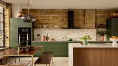 A modern kitchen with a view of the large kitchen island with marble worktop and wooden cupboards, the walls are exposed brick with green cupboards on the far wall and marble backsplashes.