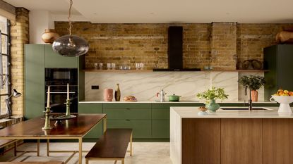 A modern kitchen with a view of the large kitchen island with marble worktop and wooden cupboards, the walls are exposed brick with green cupboards on the far wall and marble backsplashes.