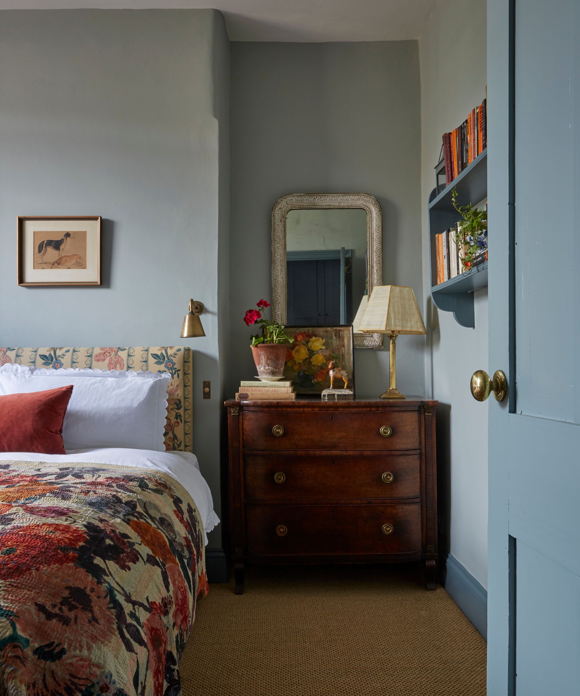 Bedroom with pale blue walls, vintage mahogany dresser, floral headboard and bedding with floral blanket, vintage prints and a blue painted bookcase