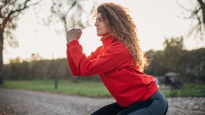 A woman in an orange jacket and leggings clasps her hands together as she executes a squat outside. Her knees and hips are bent as she moves down into the position. Behind her we can see grass, a gravel path and trees.