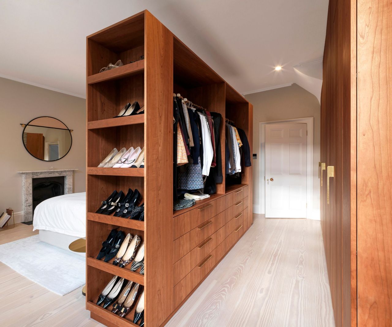 A spacious bedroom with pale wood floors and a large freestanding wooden wardrobe unit. Open shelves display neatly arranged shoes, while hanging rails hold clothing. A white bed and marble fireplace sit behind, with a round mirror above the mantel.