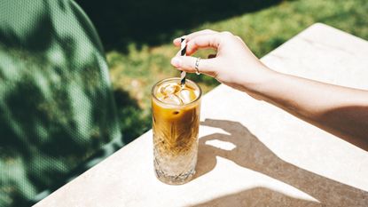 Is cold brew the same as iced coffee? Here is a glass cup of brown iced coffee on a light wooden surface, with a hand reaching over to put in a black and white straw, with a lawn underneath the surface