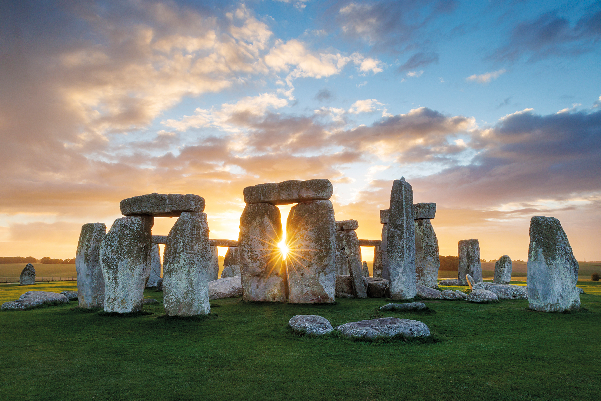 Sunset shining through the ancient stone circle of Stonehenge with a partly cloudy sky and green grass foreground