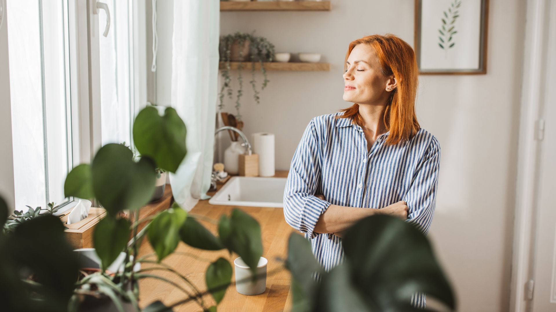 Woman standing in kitchen with plants, looking towards open window with closed eyes and arms folded, representing symptoms of perimenopause