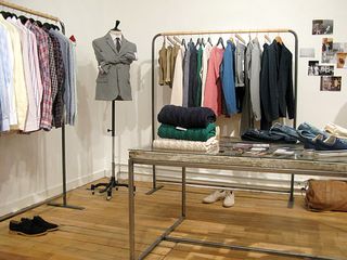 A collection of shirts on the left rail, a mannequin in the corner with a grey suit jacket, a collection of leisure clothing, a table with three cable knit jumpers, trainers and jeans.