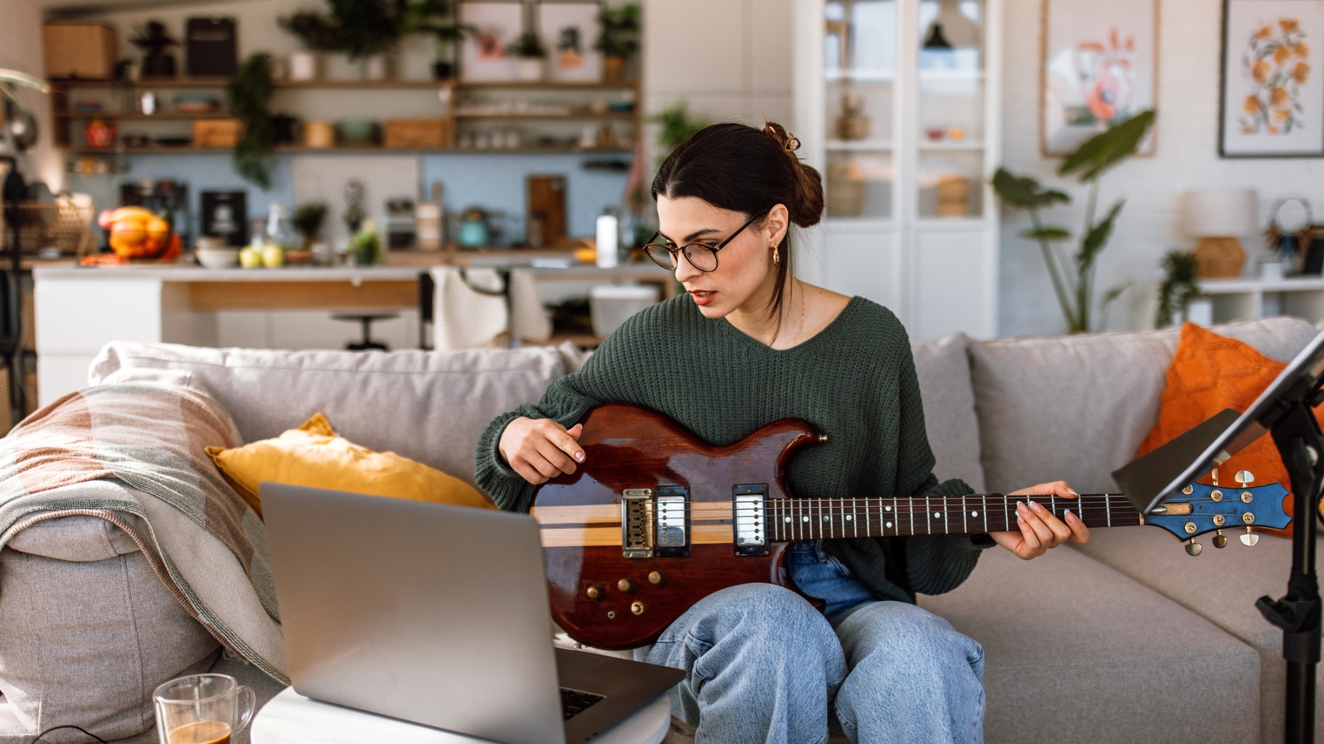 A woman sits in front of a laptop on a grey sofa, with an electric guitar in her lap