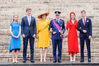 Princess Eléonore in a blue dress, Prince Gabriel in a blue suit, Queen Mathilde in a yellow dress, King Philippe in a blue military uniform, Princess Elisabeth in a red dress and Prince Emmanuel in a blue suit standing at the bottom of a staircase