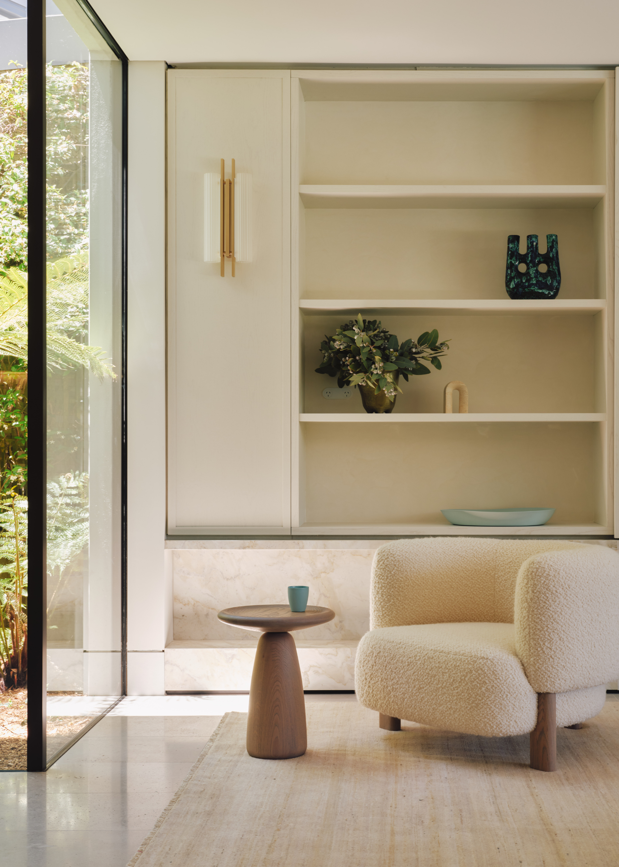 A living room with a bright window and a boucle white armchair beside a wood side table against a shelf of vases and bowls
