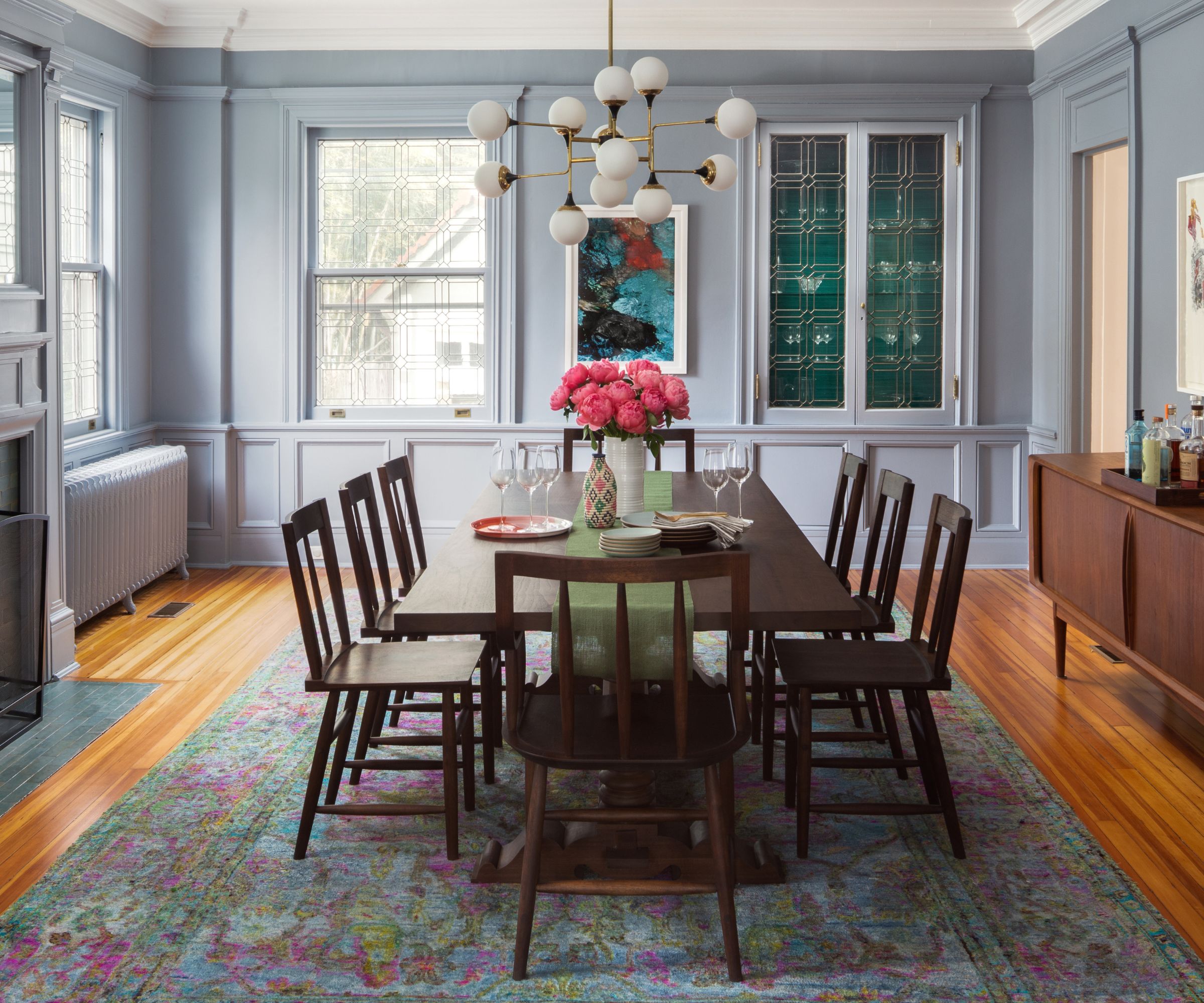 A dining room with gray color-drenched walls, a large dark wooden dining table and chairs, a jewel-toned area rug, and teal artwork on the walls.