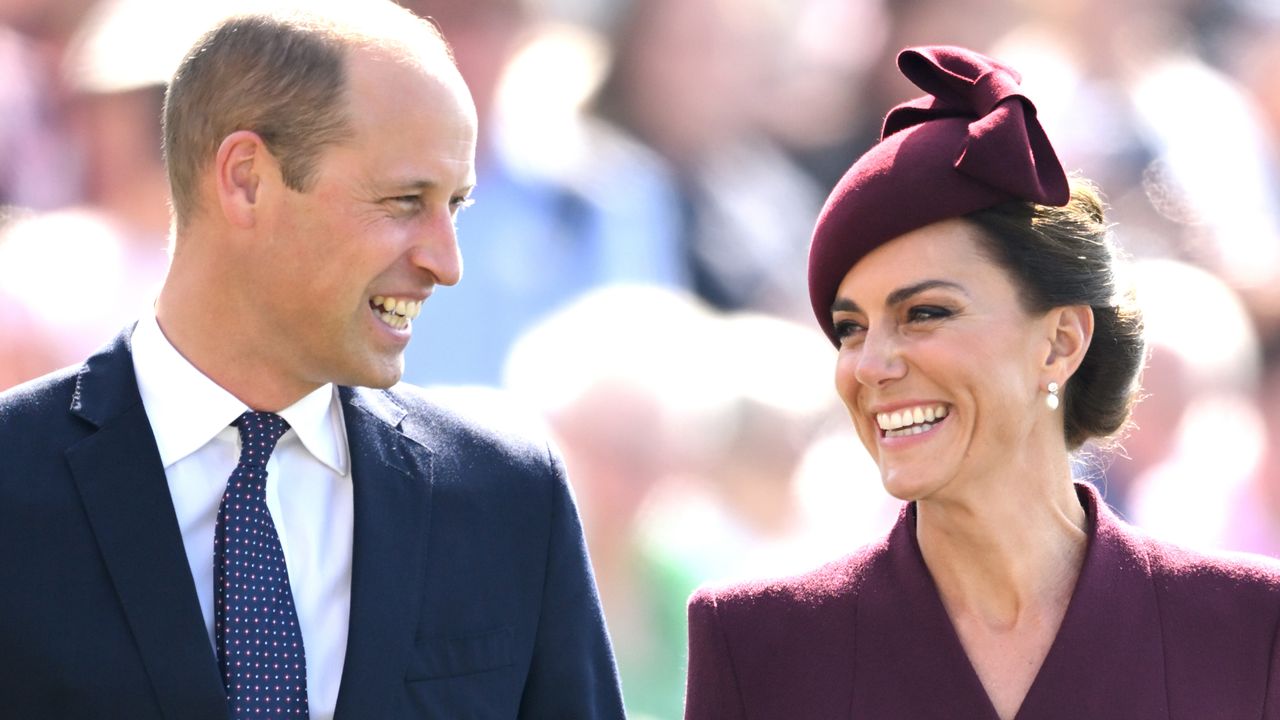 Kate Middleton wearing a purple coat and hat and Prince William wearing a suit smiling at each other 