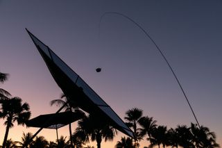 Dusk image, silhouette palm trees, close up of a solar reflector and one in the distance, extended carbon rod dangling a small dark object above the centre reflector