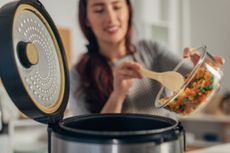 Woman using a wooden spoon to empty a glass bowl of chopped veg into slow cooker
