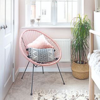 Grey carpet with fuffly rug, pink chair and cushions next to freestanding potted plant in wicker basket