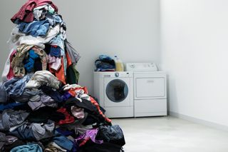 A huge pile of clothes is waiting to go in the washing machine, which is in the background of a white room next to a tumble dryer