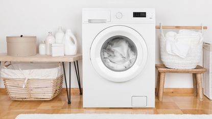 A white front-loading washing machine against a white wall, wooden floor, Wooden tables beside with white laundry products on them, and a white woven laundry basket of white linens.