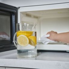 Jug of water with lemons in it standing in front of a microwave while someone uses a white cloth to clean the inside of the microwave