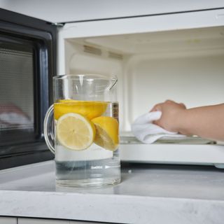 Jug of water with lemons in it standing in front of a microwave while someone uses a white cloth to clean the inside of the microwave