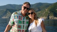 Alan Carr and Amanda Holden smile and pose with each other with Italian mountains and water behind them.