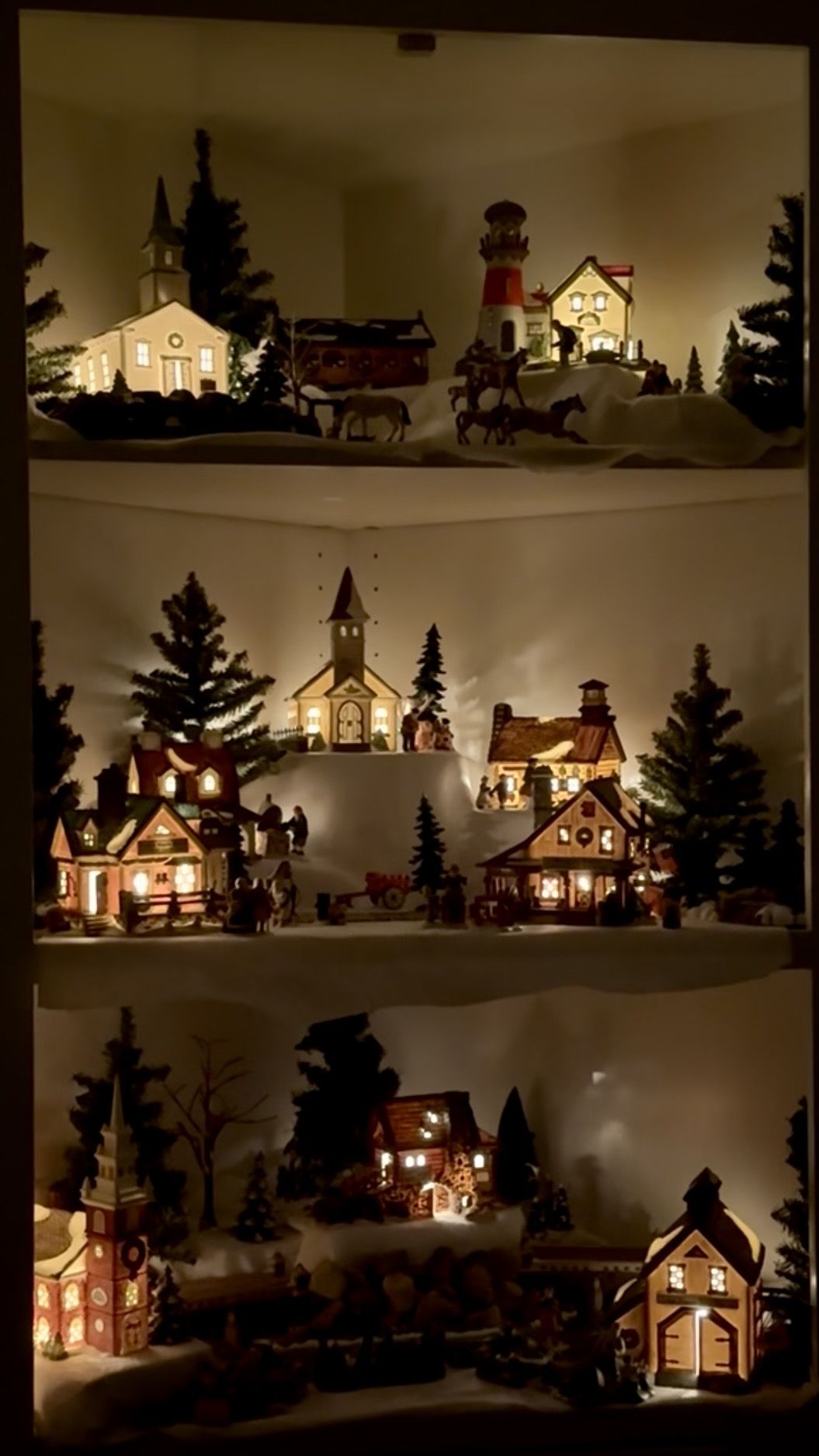 Christmas village displays inside open corner shelving with lit miniature houses, trees fake snow and mini animals and people with the main house lights turned off so only the mini houses are glowing