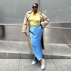 Editor Aniyah Morinia in blue satin skirt and yellow polo top with trench coat and blue ballet flats.