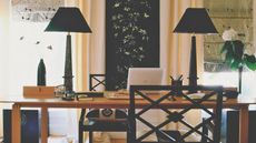 Home office space with black lamps, black chairs, and a large wooden desk with a laptop and pens. To either side are bright windows with patterned blinds, flowers, and vases.