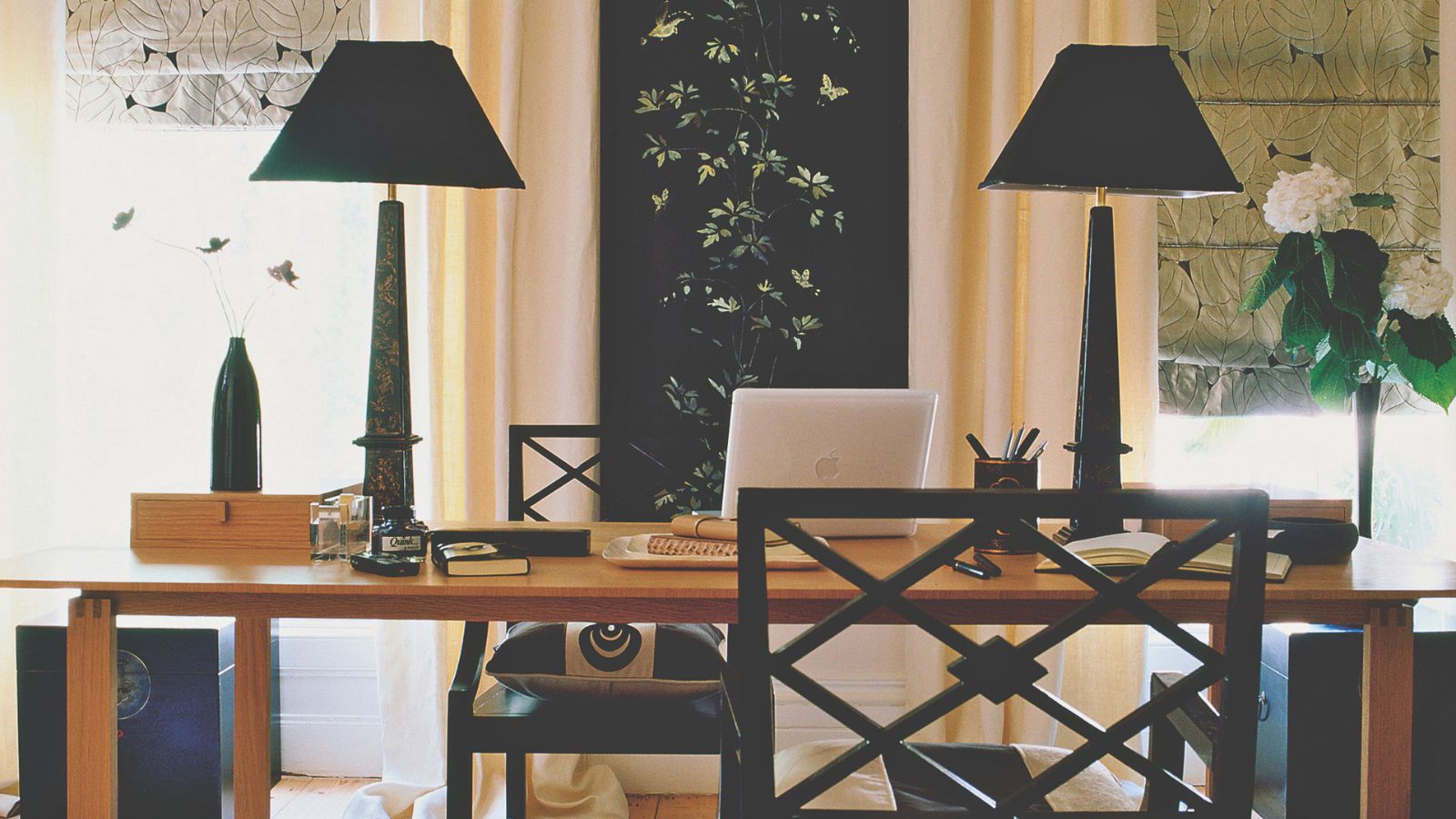 Home office space with black lamps, black chairs, and a large wooden desk with a laptop and pens. To either side are bright windows with patterned blinds, flowers, and vases.