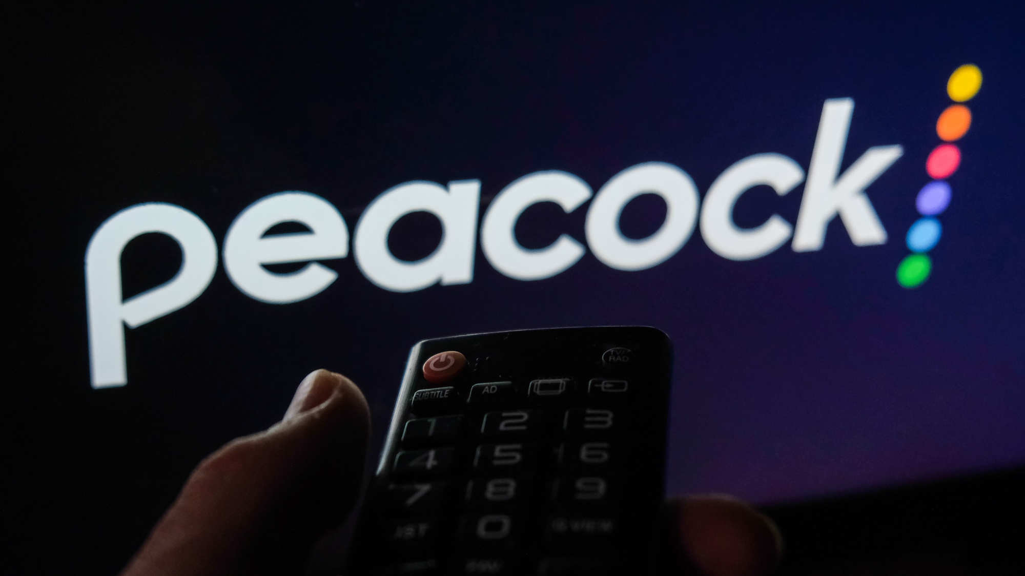 Control remoto sostenido frente a la pantalla con el logotipo de Peacock TV