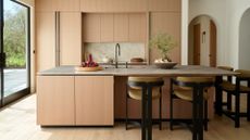 A bright minimalist kitchen with wood cabinets, a marble backsplash, a wood island with a stone top, a mini potted tree, a bowl of plums, and four chairs