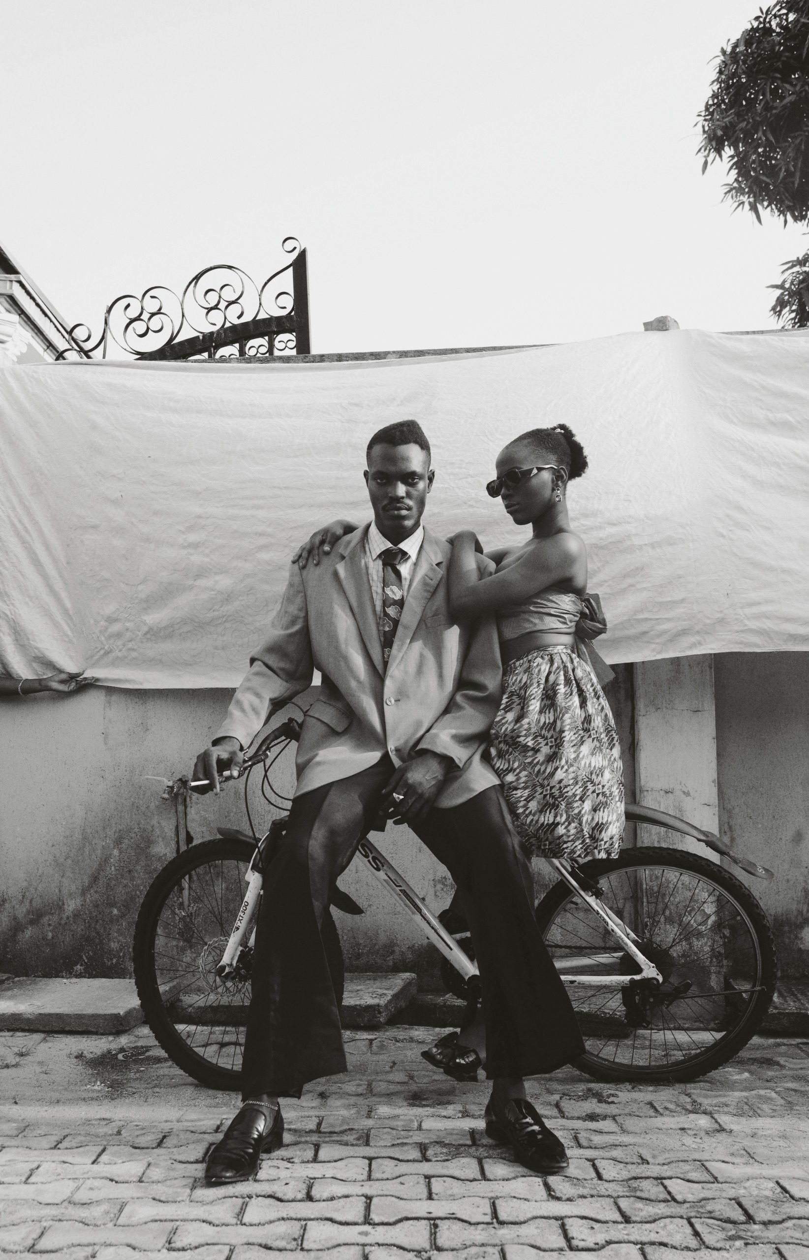 A black and white portrait of a man in a suit and a woman in a patterned skirt posing together on a bicycle against a draped white backdrop.