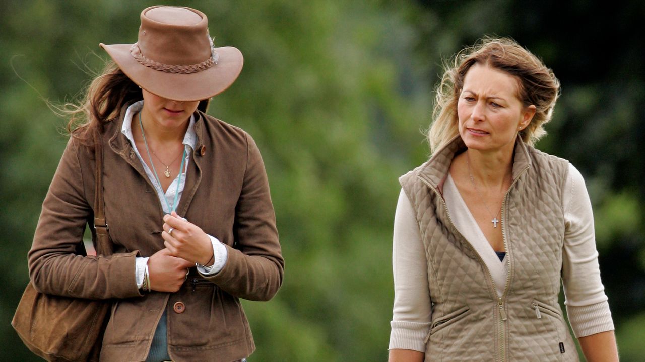 Kate Middleton wearing brown jacket and cowboy hat and walking next to Carole Middleton wearing a tan vest in front of green trees 