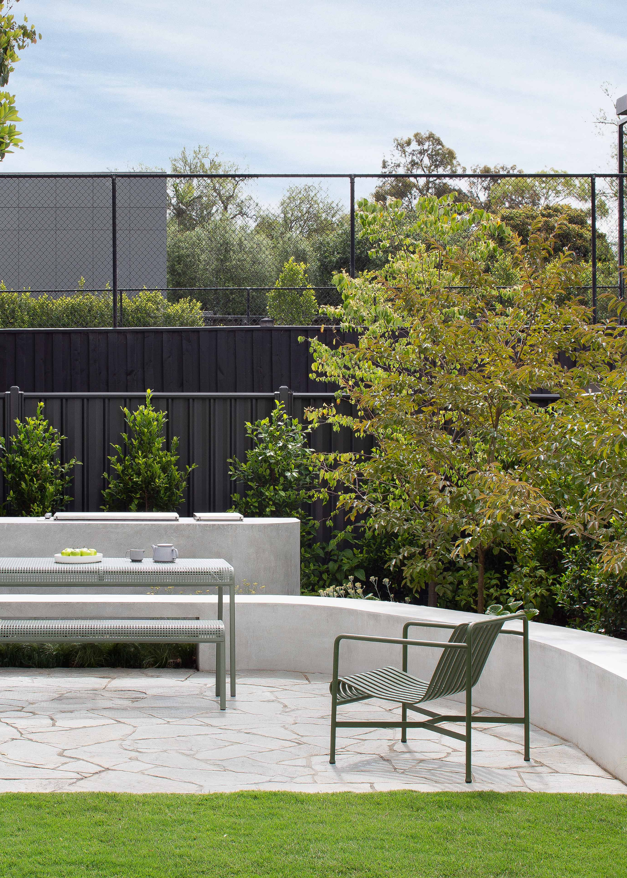 A backyard with metal green outdoor furniture, tall trees and a black fence