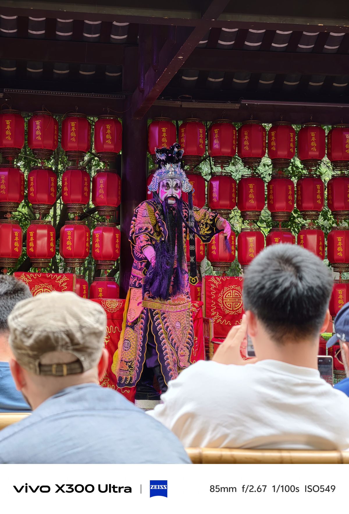 An actor in an ornate purple costume performing face-changing in front of red lanterns.
