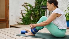 woman in turquoise leggings and white tshirt, sitting on an exercise mat strapping on an ankle weight around her ankle. She's on a wooden outdoor floor.