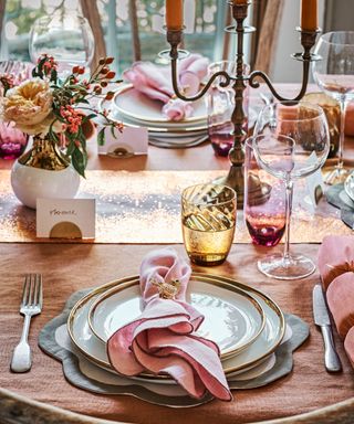 Thanksgiving centerpiece ideas with pink napkins, gold printed runner, candlestick with orange candles and gold and white ceramic vases