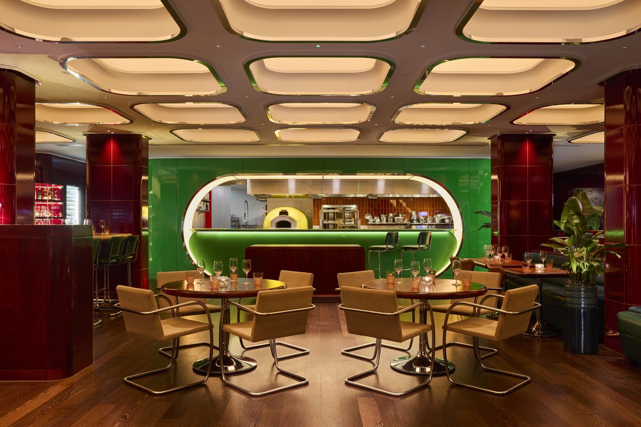 The interiors of a fine dining restaurant feature coffered, back-lit ceilings with chrome detailing, cherry plum walls covered in glazed ceramic, dark green banquettes, and atmospheric sconces, as well as plants.