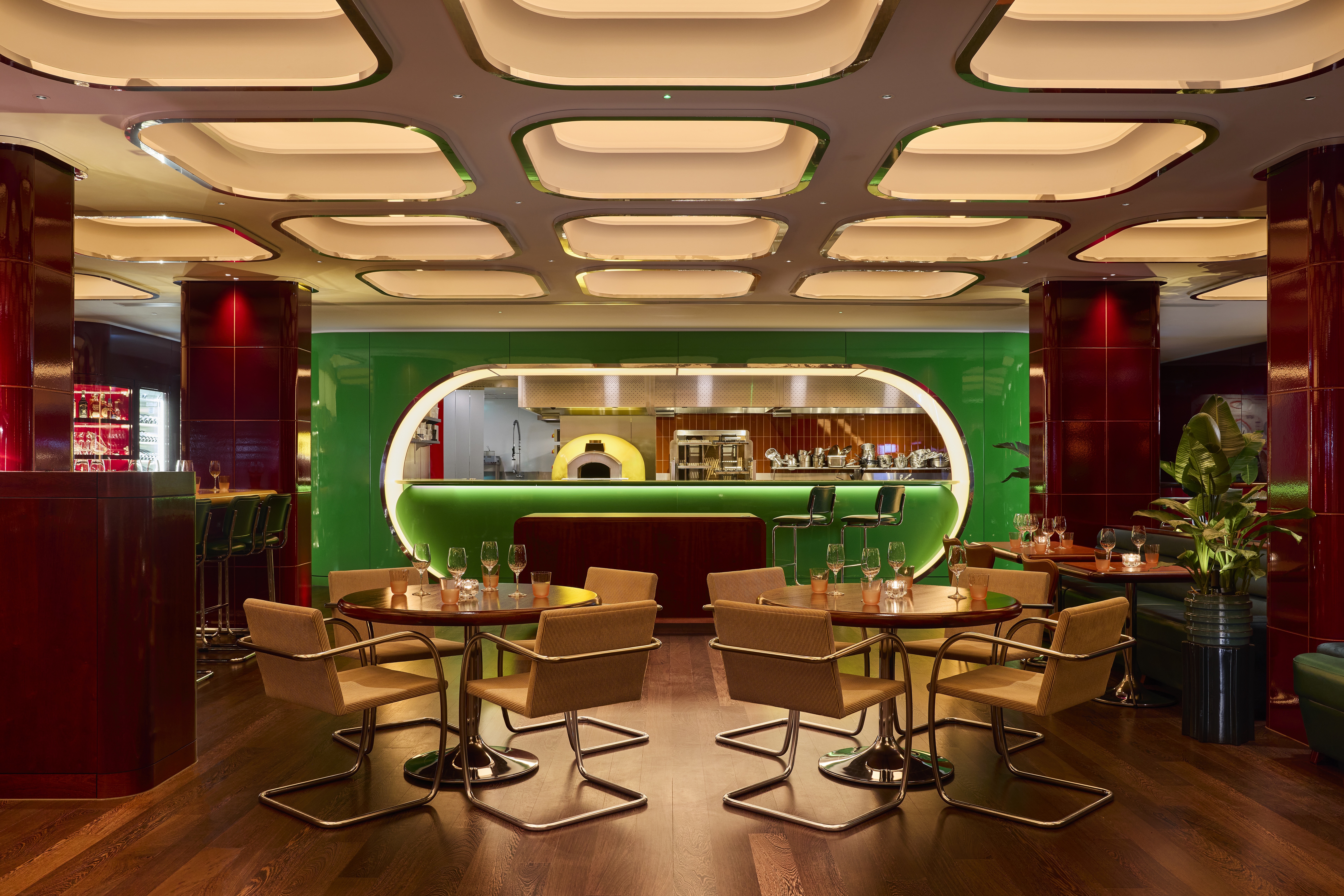 The interiors of a fine dining restaurant feature coffered, back-lit ceilings with chrome detailing, cherry plum walls covered in glazed ceramic, dark green banquettes, and atmospheric sconces, as well as plants.