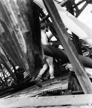 Close-up black and white photo of man in work boots and denim shorts seen through pier structure