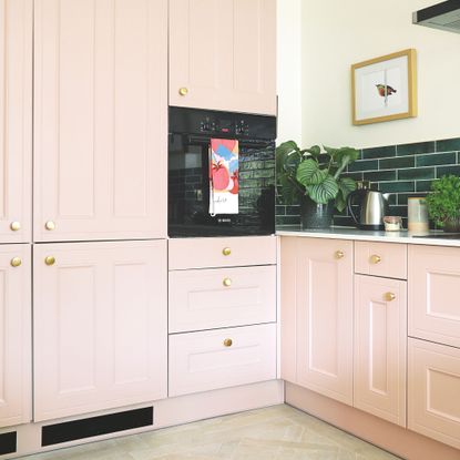 Kitchen with pink cabinetry, green tiles on the wall, and white countertops. There are black plinth heaters above the tiled floor